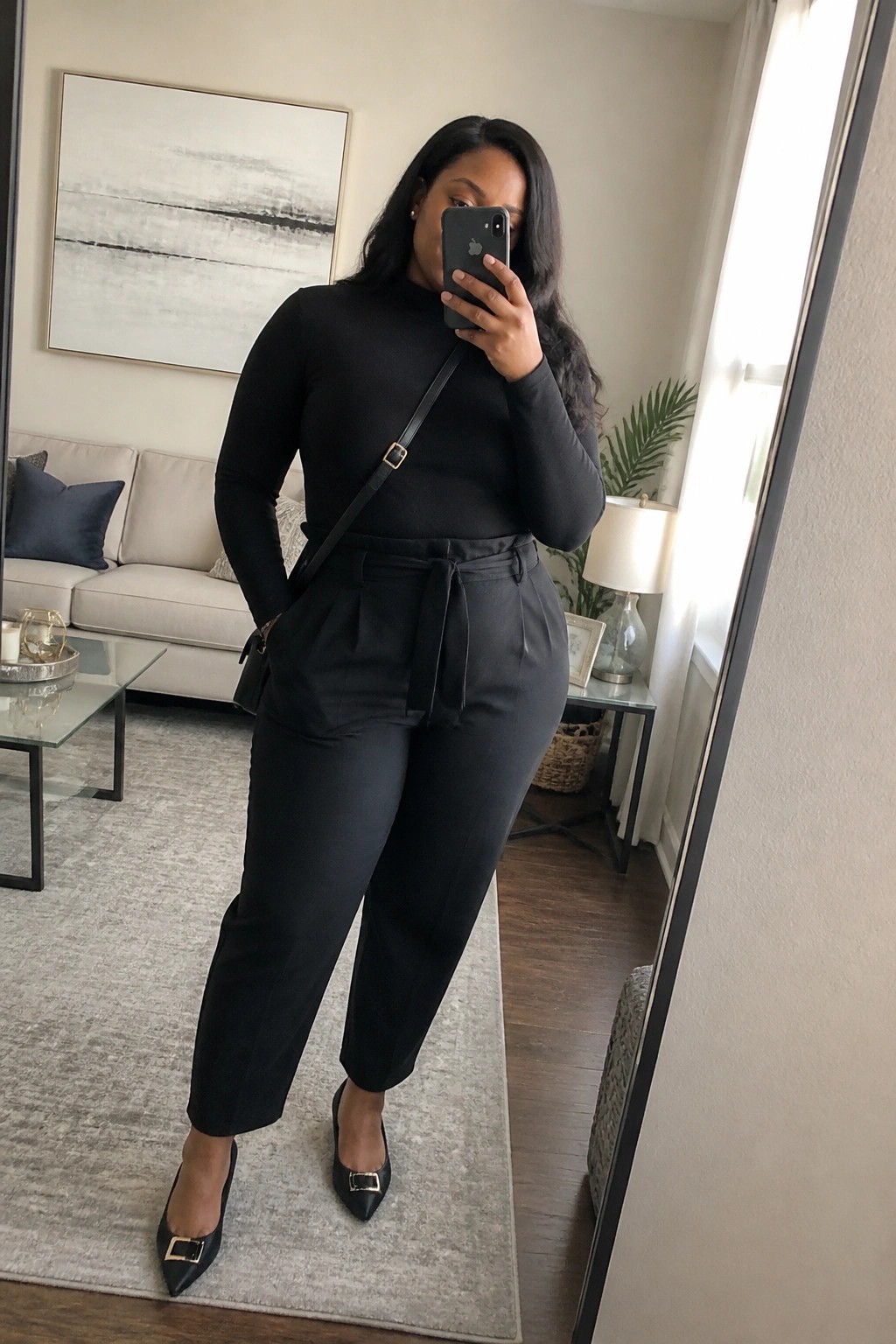 A plus size Black woman in a mirror selfie wearing an all black turtleneck top tucked into high waisted belted trousers with pointed toe flats and a small crossbody bag