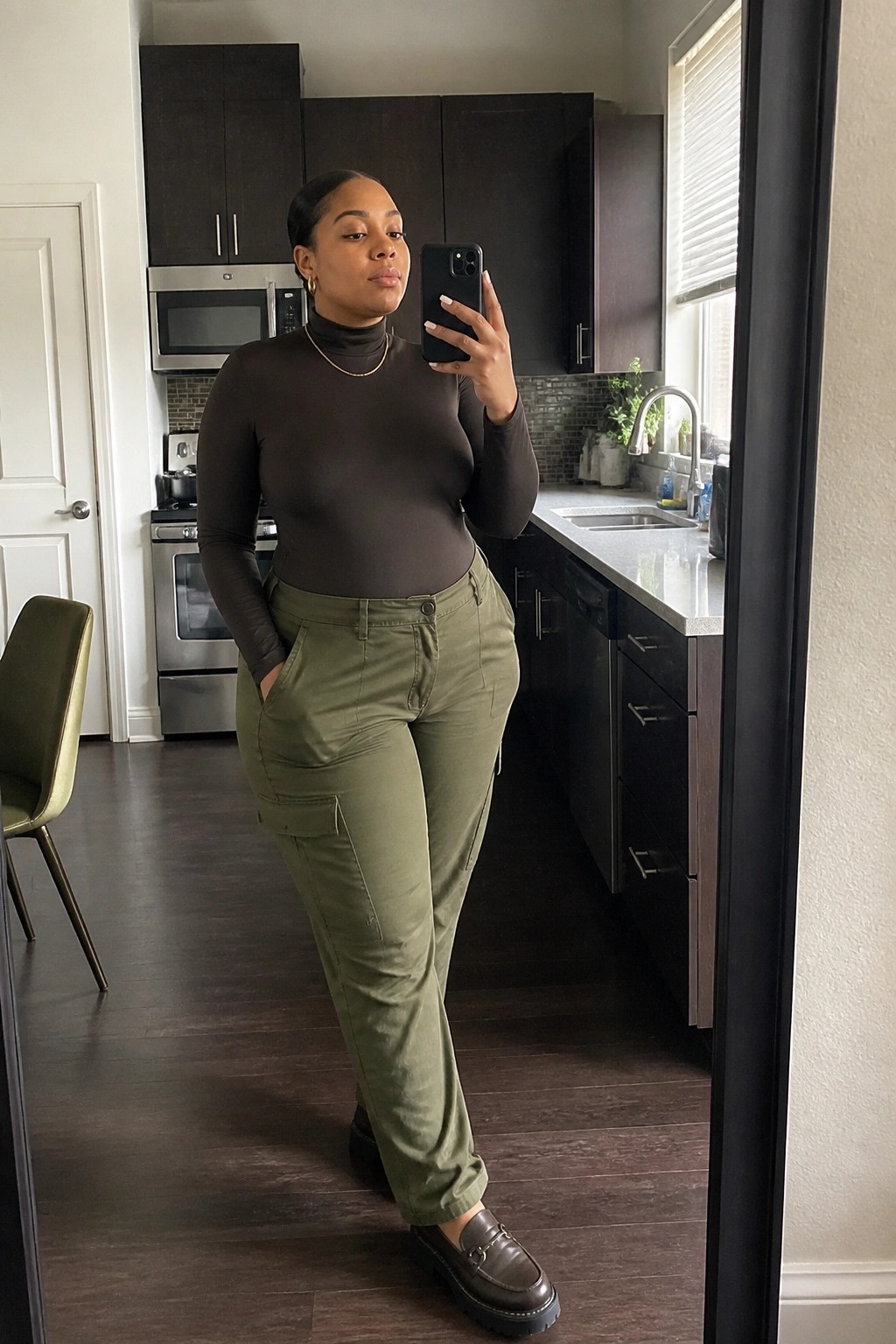 Woman wearing a fitted brown turtleneck bodysuit tucked into olive green cargo pants with side pockets, paired with dark brown loafers and a thin gold necklace, standing in a kitchen for a mirror selfie.