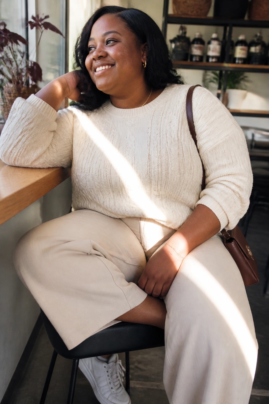 Smiling woman in cream cable-knit sweater and beige pants in sunlit cafe