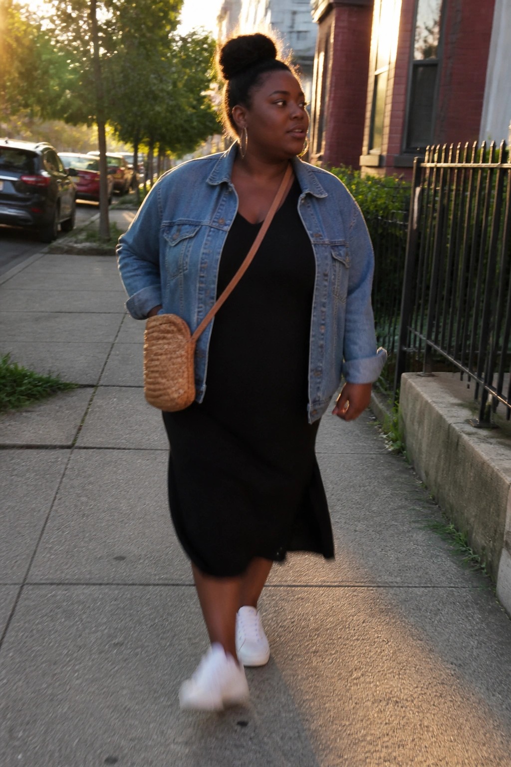 Woman in denim jacket and black dress walks on sunny urban sidewalk with woven bag.
