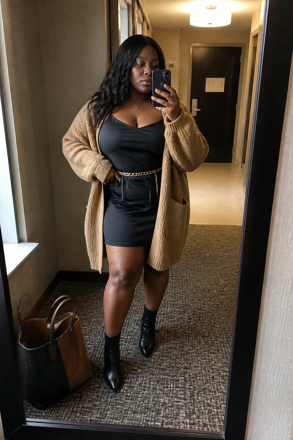 Plus size Black woman in a black satin mini dress layered with an oversized beige fuzzy cardigan, gold chain belt at the waist, black ankle boots, holding a phone for a mirror selfie in a hallway.