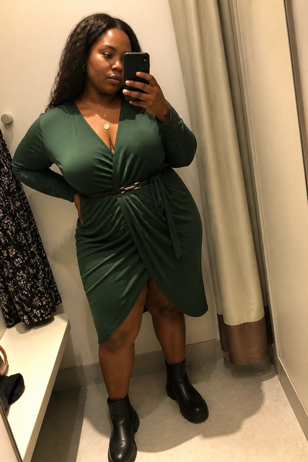 Woman in green wrap dress and black boots taking mirror selfie in fitting room