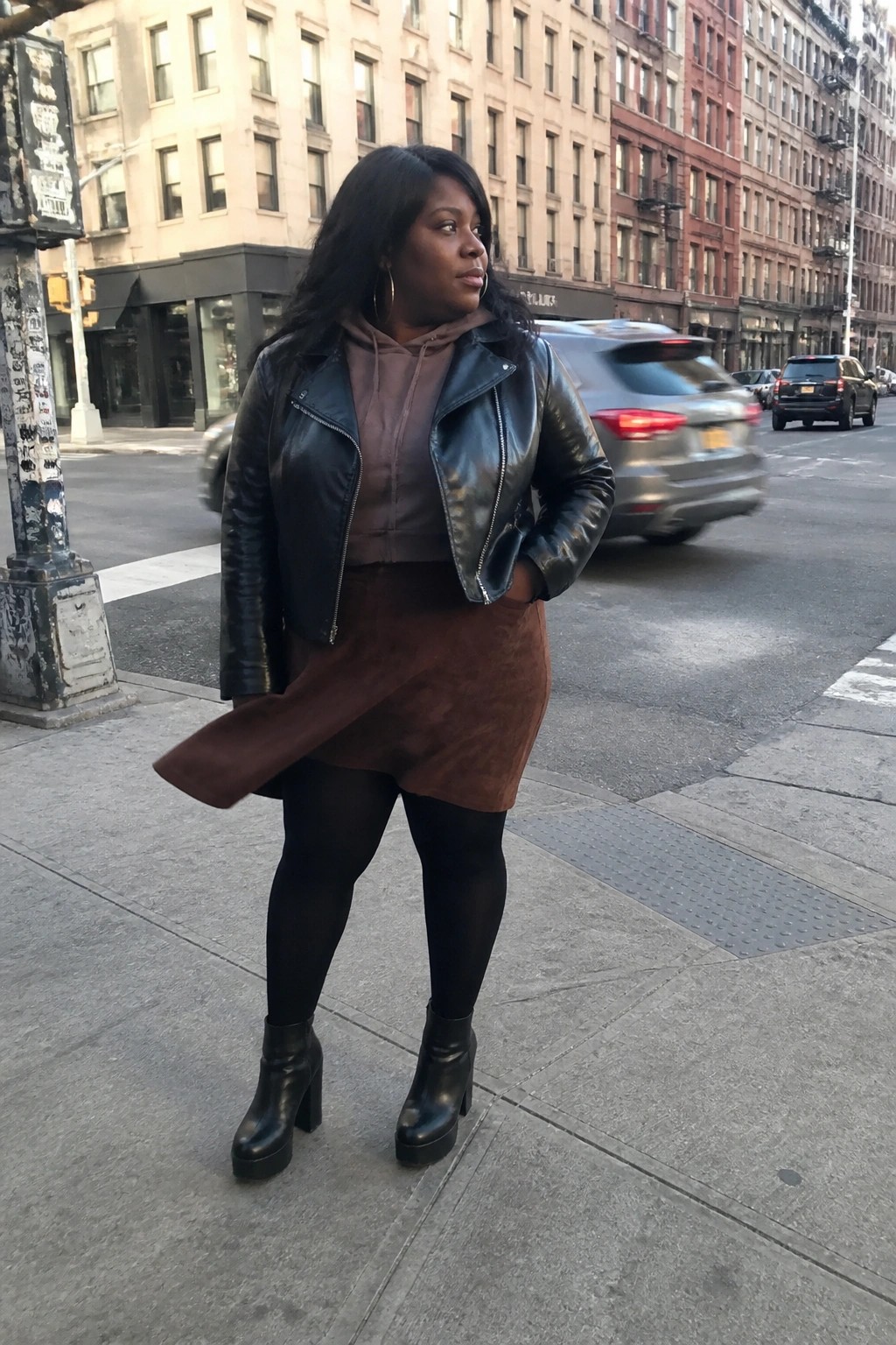 Woman in black leather jacket and brown suede skirt on city sidewalk.