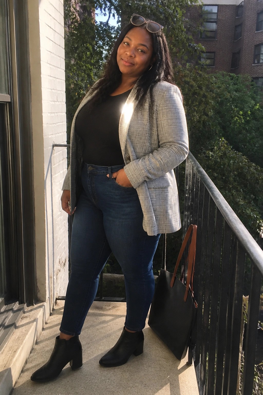 Woman in plaid blazer and jeans on sunny balcony with tote bag and sunglasses