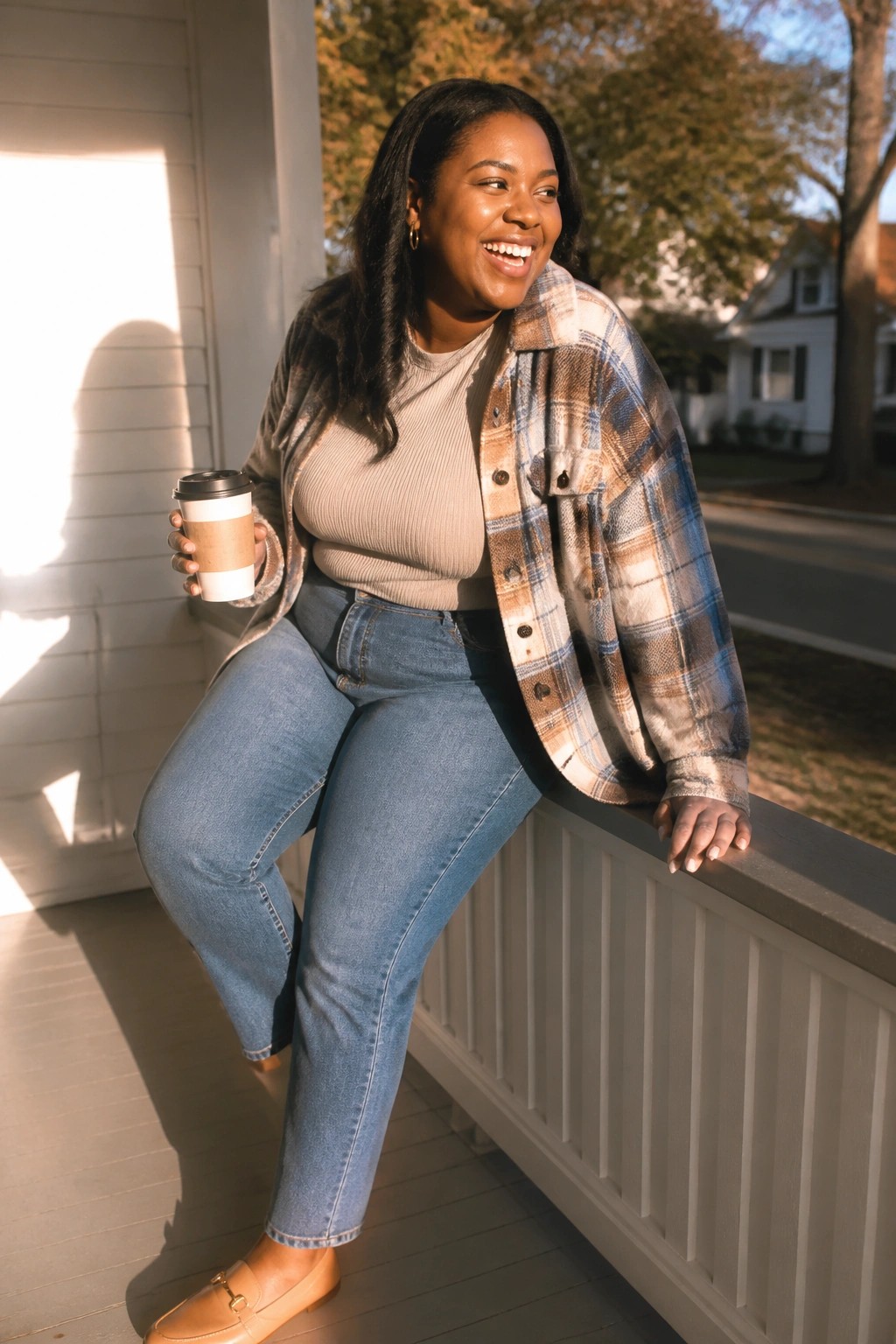 Smiling woman in plaid shirt and jeans holding coffee on sunny porch railing