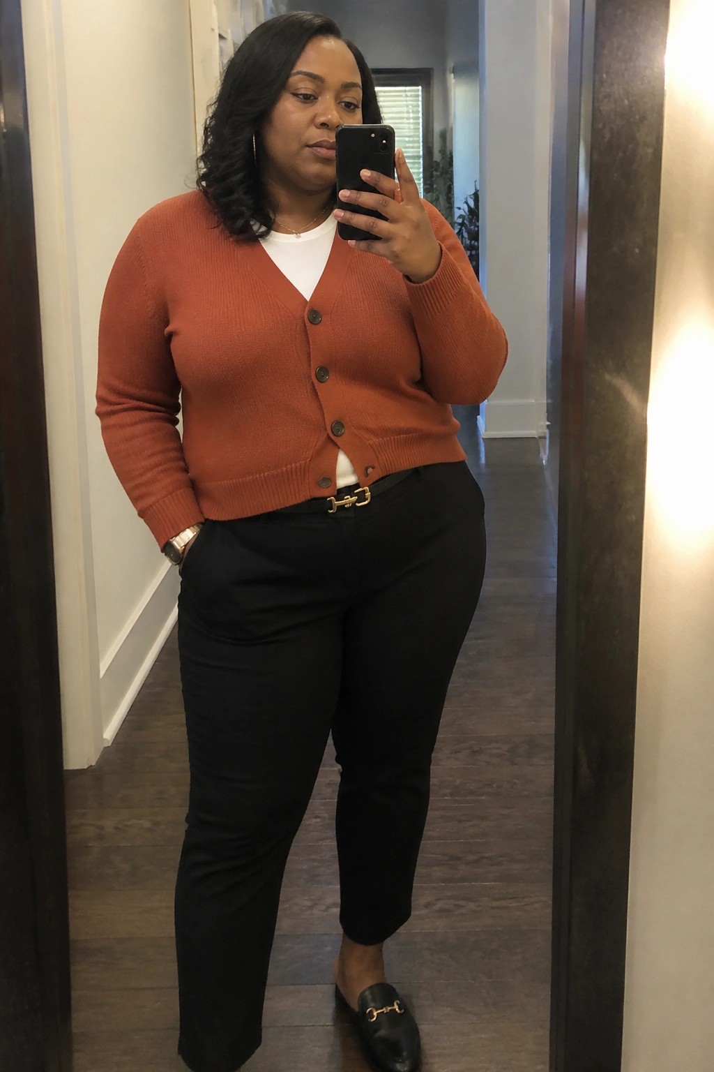 Plus size Black woman in mirror selfie wearing rust orange button cardigan over white tee black slim pants with gold buckle belt and black loafers with gold trim.
