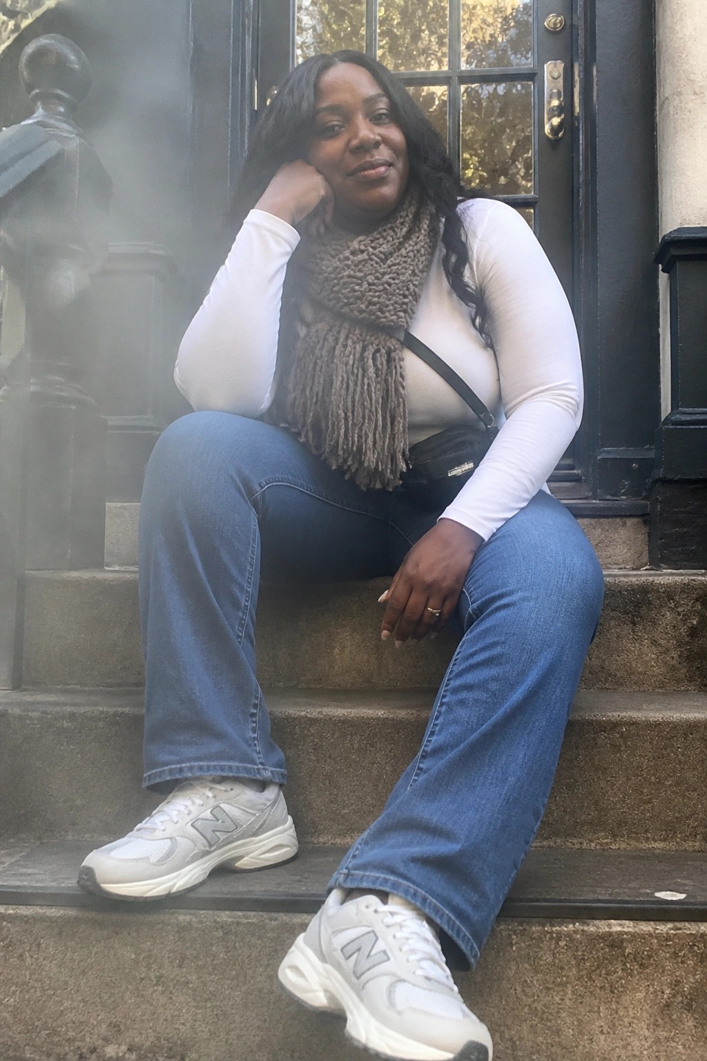 Woman in white top, blue jeans, brown scarf sits on stone steps by door.