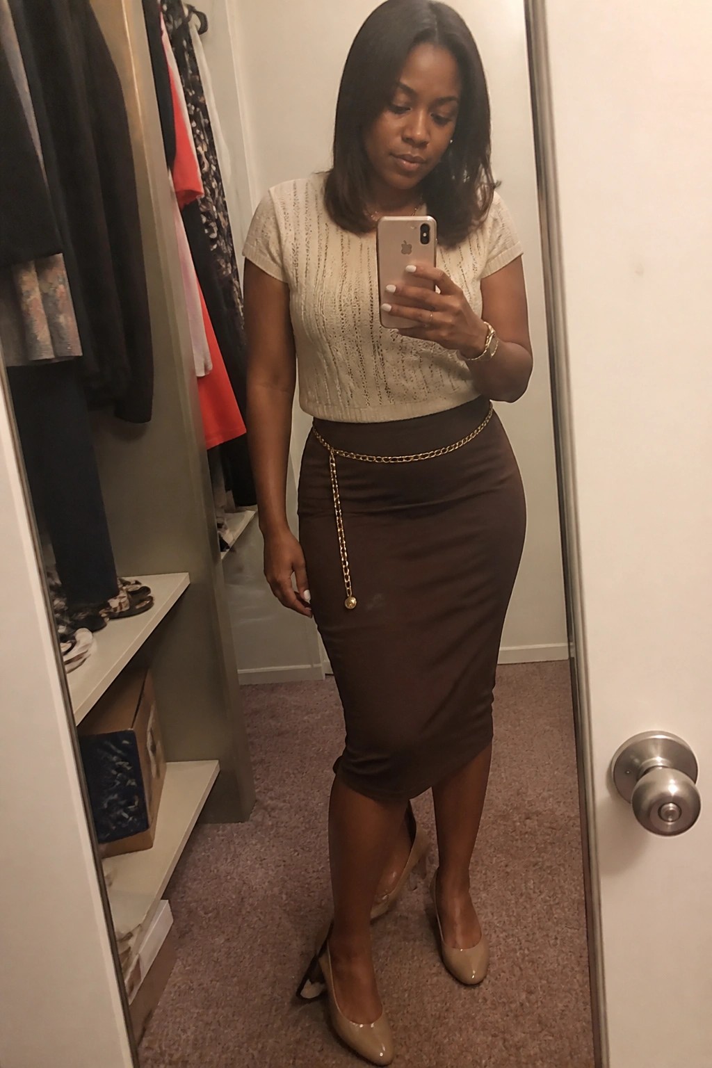 Woman wearing a cream textured knit short sleeve top with a brown midi pencil skirt accented by a gold chain belt and beige pointed toe pumps while taking a mirror selfie.