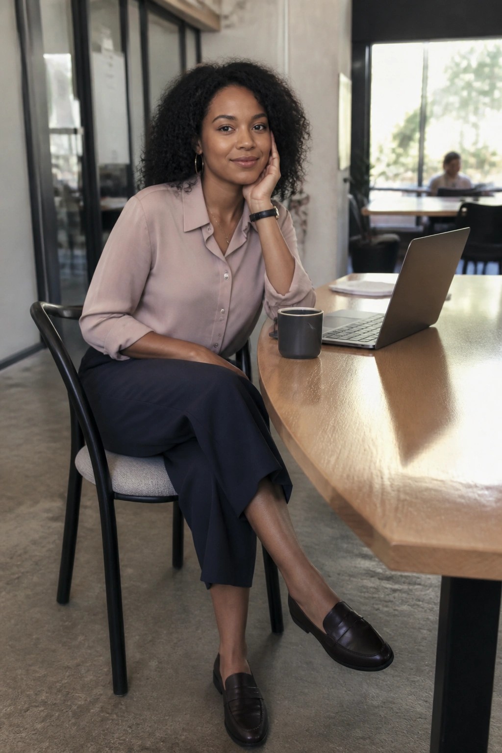 Curly-haired woman in pink blouse smiling at wooden table with laptop and mug
