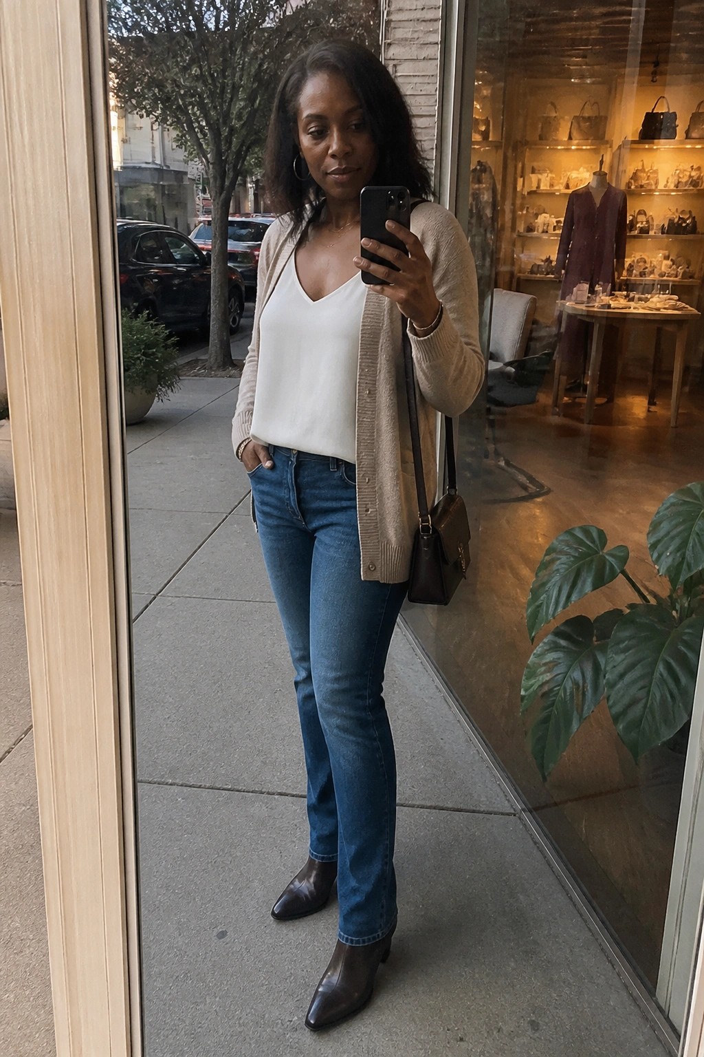 Woman in white top, beige cardigan, and jeans taking mirror selfie outside boutique