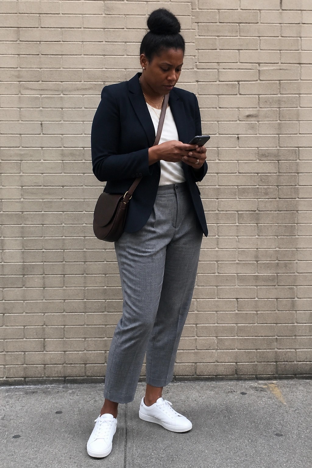 Woman in navy blazer and gray pants using phone by brick wall