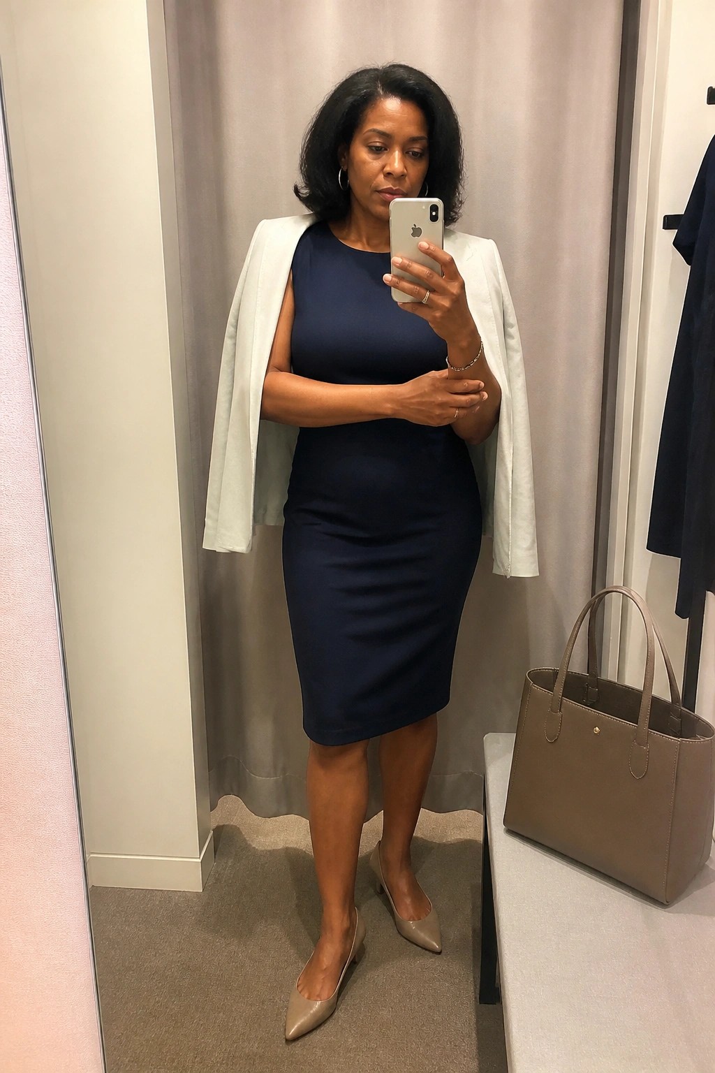 Woman wearing a navy blue sleeveless sheath dress with a white blazer draped over her shoulders, beige pointed toe heels, and holding a smartphone in a fitting room setting with a taupe handbag nearby.