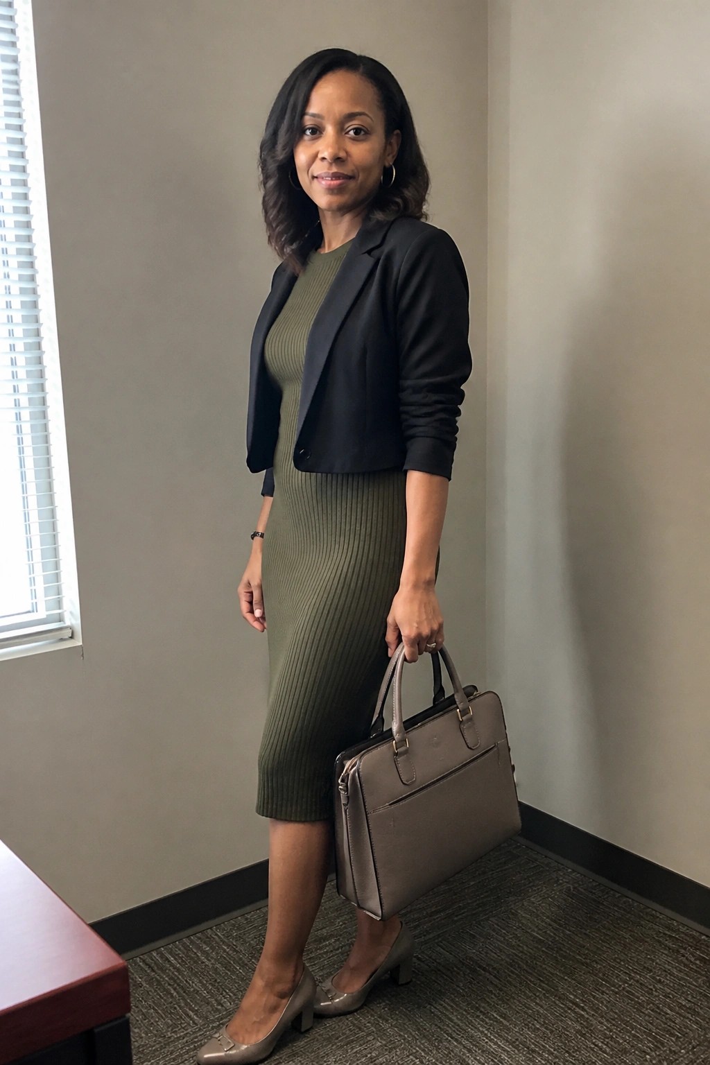 Woman in olive ribbed dress and black cropped blazer holding briefcase in office.