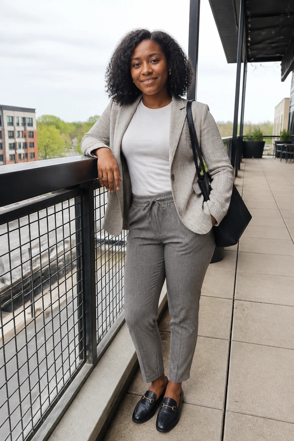 A woman in a light gray plaid blazer over a white ribbed top, gray drawstring pants, black loafers with gold hardware, and a black tote bag.