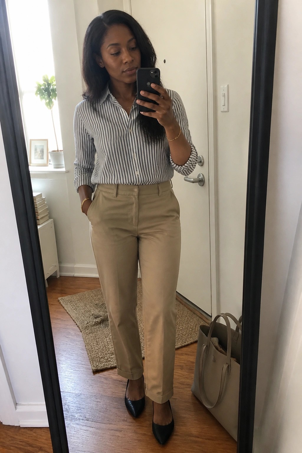 Black woman in a black and white striped button down shirt tucked into beige khaki trousers paired with black pointed toe heels and gold jewelry taking a mirror selfie.