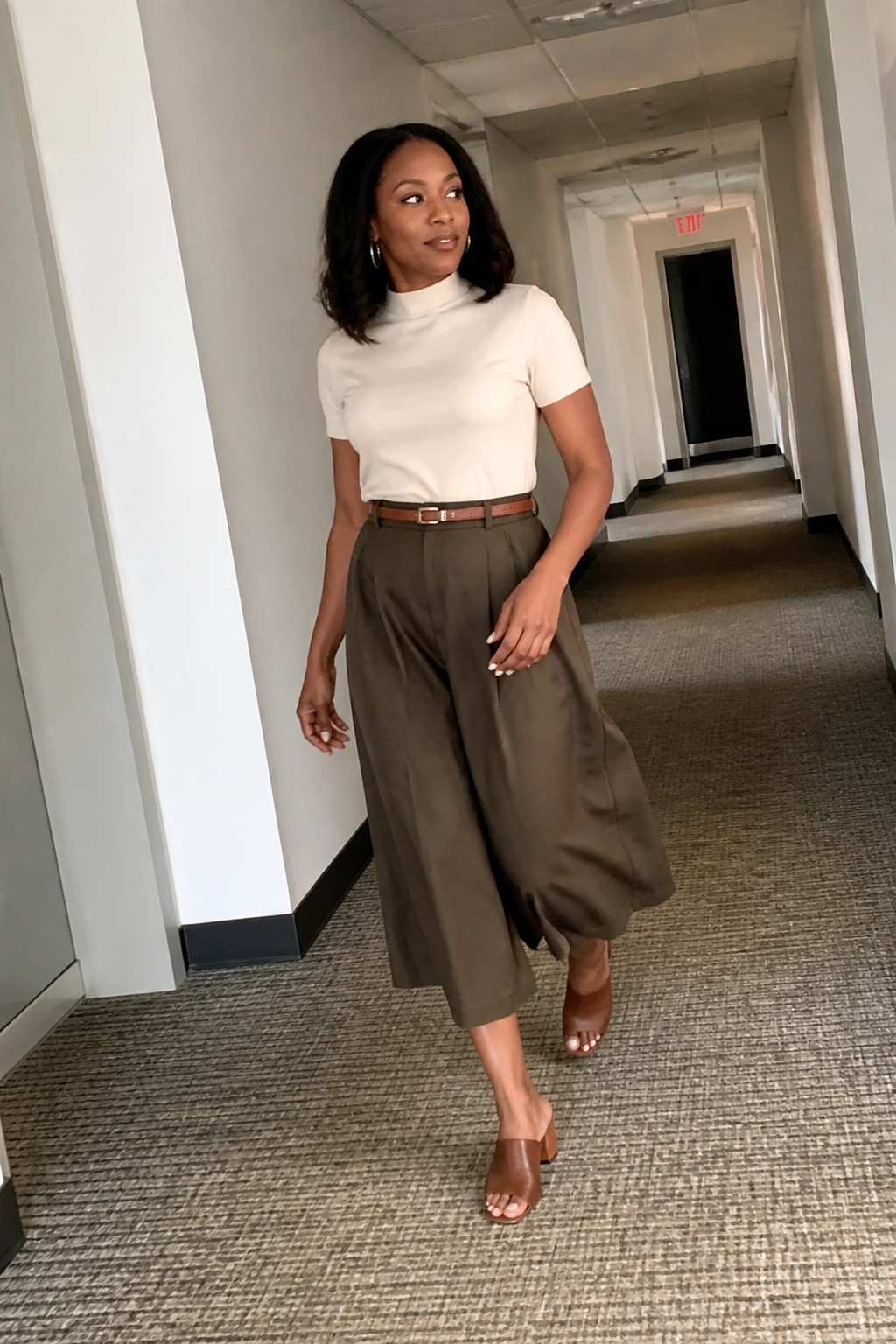 Woman in white turtleneck and olive wide-leg pants posing in hallway