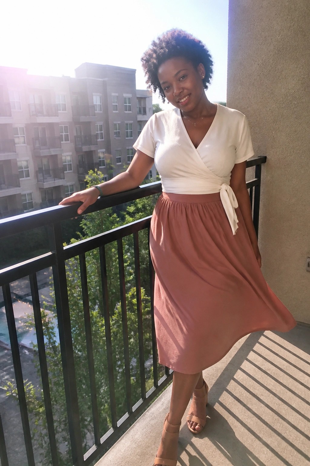 Woman with curly hair in white wrap top and pink skirt on sunny balcony