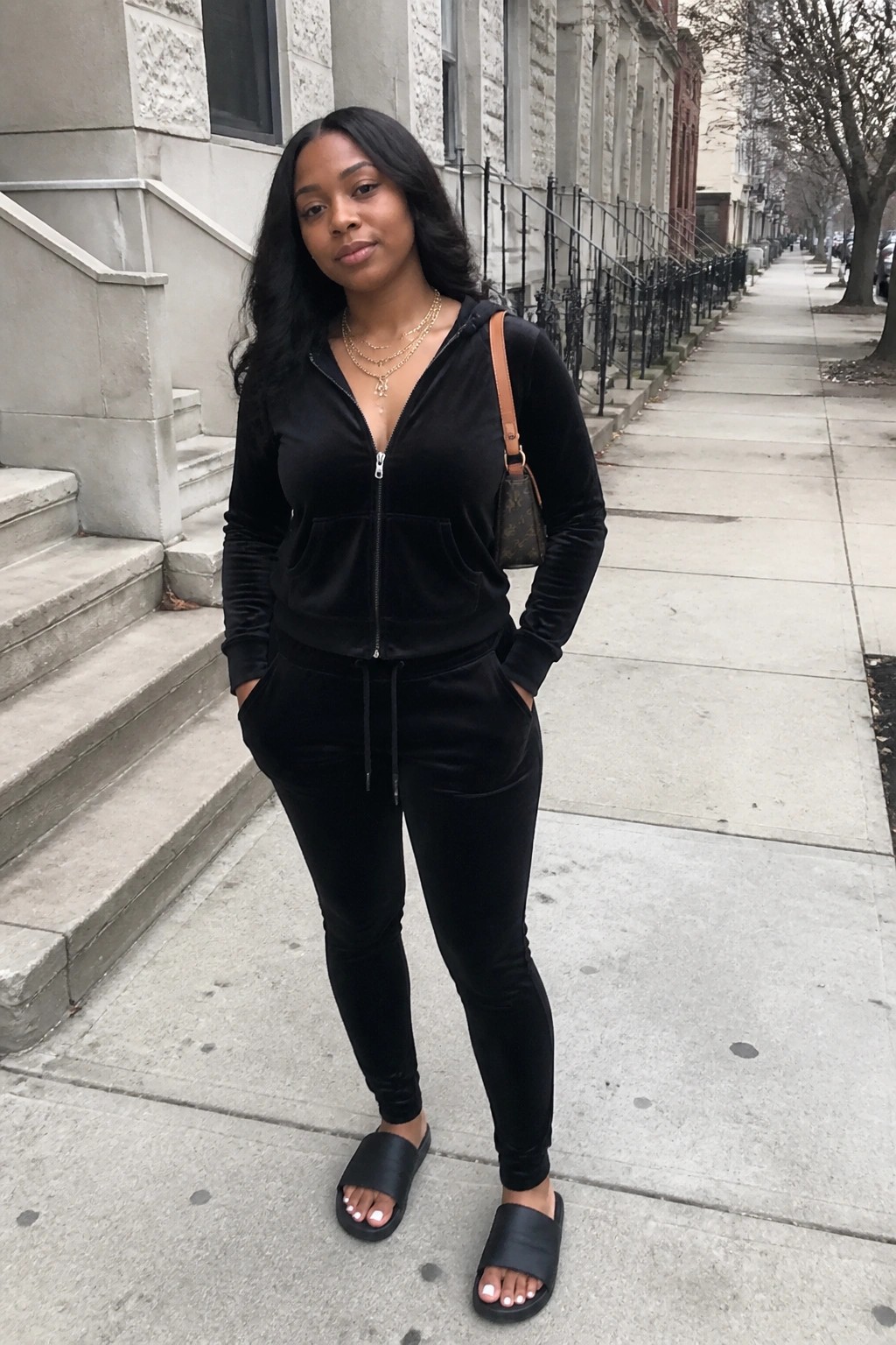 Woman in black velvet tracksuit and slides on urban sidewalk.
