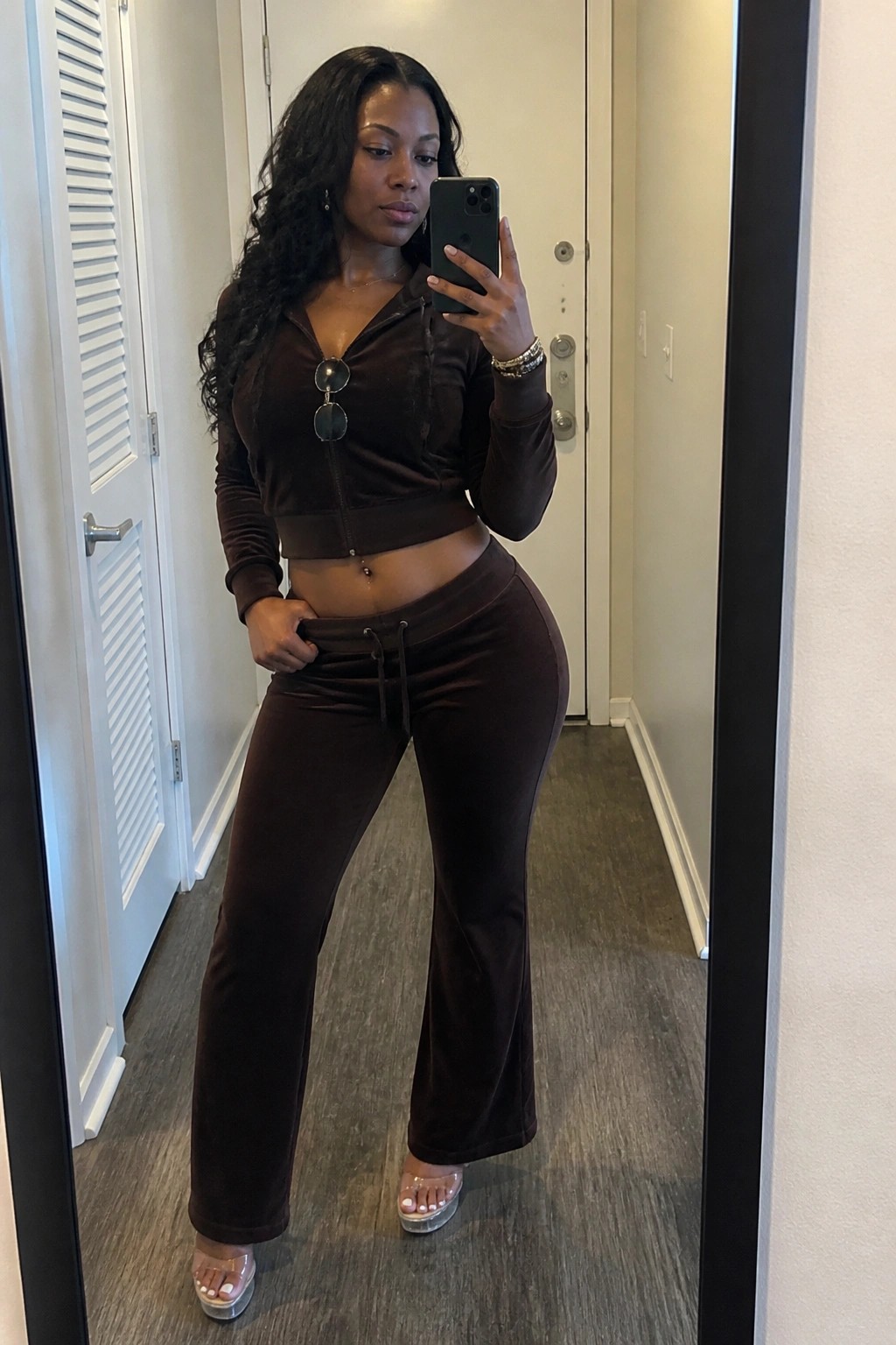 Woman in brown velour tracksuit taking mirror selfie while holding smartphone in hallway