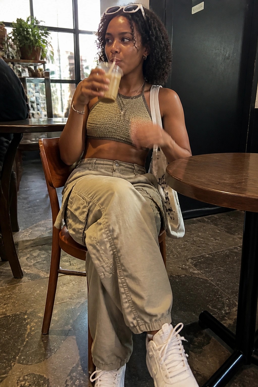 Curly-haired woman in crocheted top and cargo pants sipping beverage in cafe