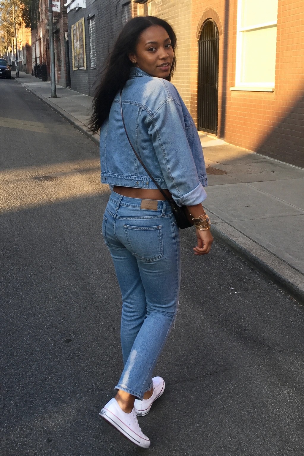 Woman in cropped light-wash denim jacket and jeans on urban sidewalk, glancing back.