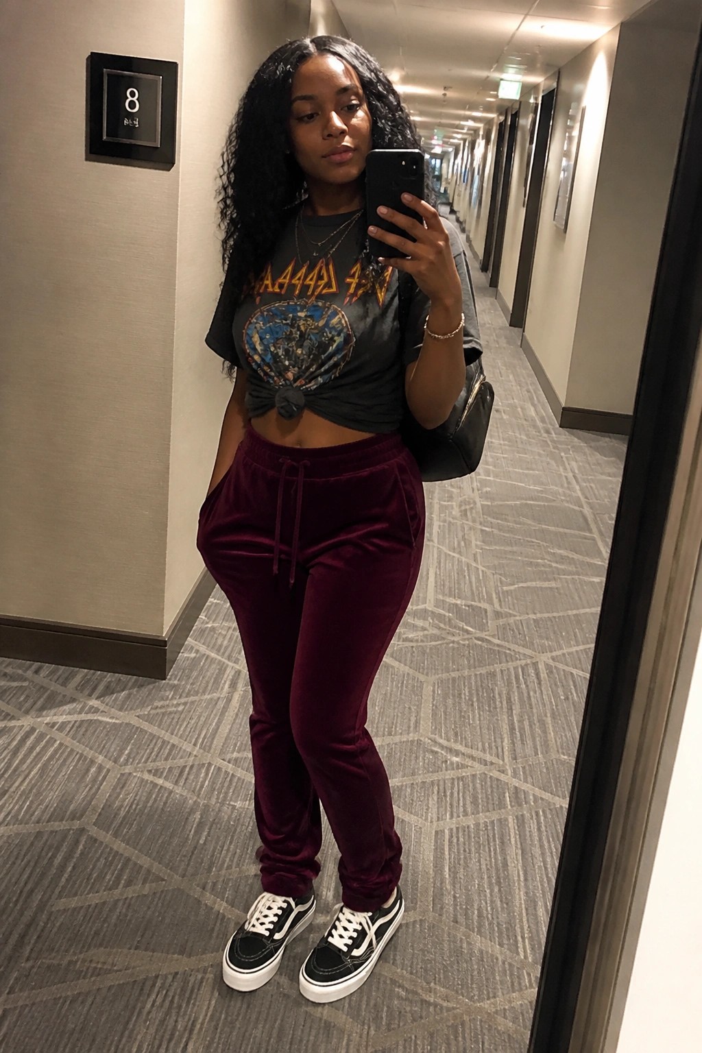 Black woman in hallway mirror selfie wearing black graphic band tee knotted at waist maroon velvet joggers and black white sneakers with layered necklaces and shoulder bag.