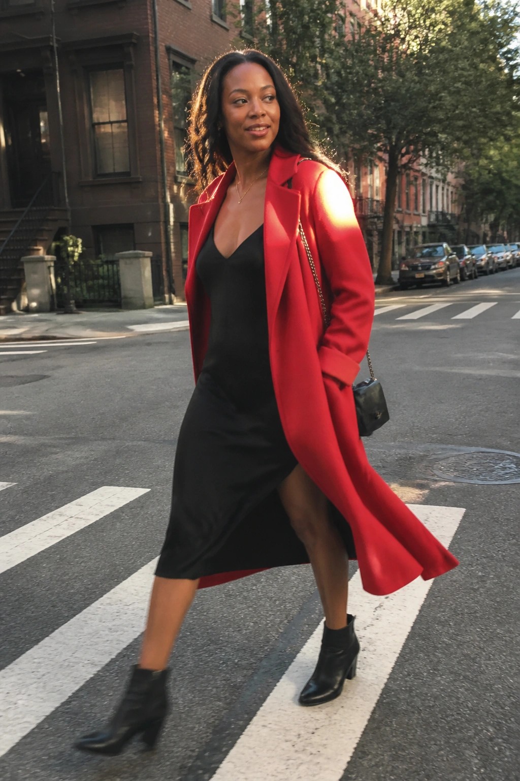 Woman in red coat over black dress crossing sunny city street with handbag