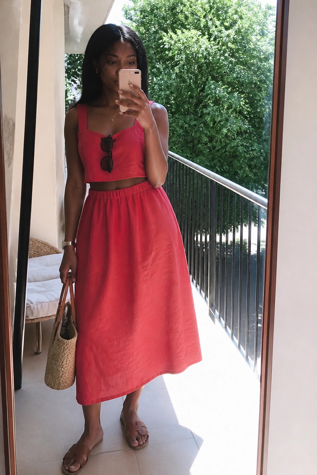 Woman in red crop top and midi skirt taking mirror selfie on balcony with straw bag
