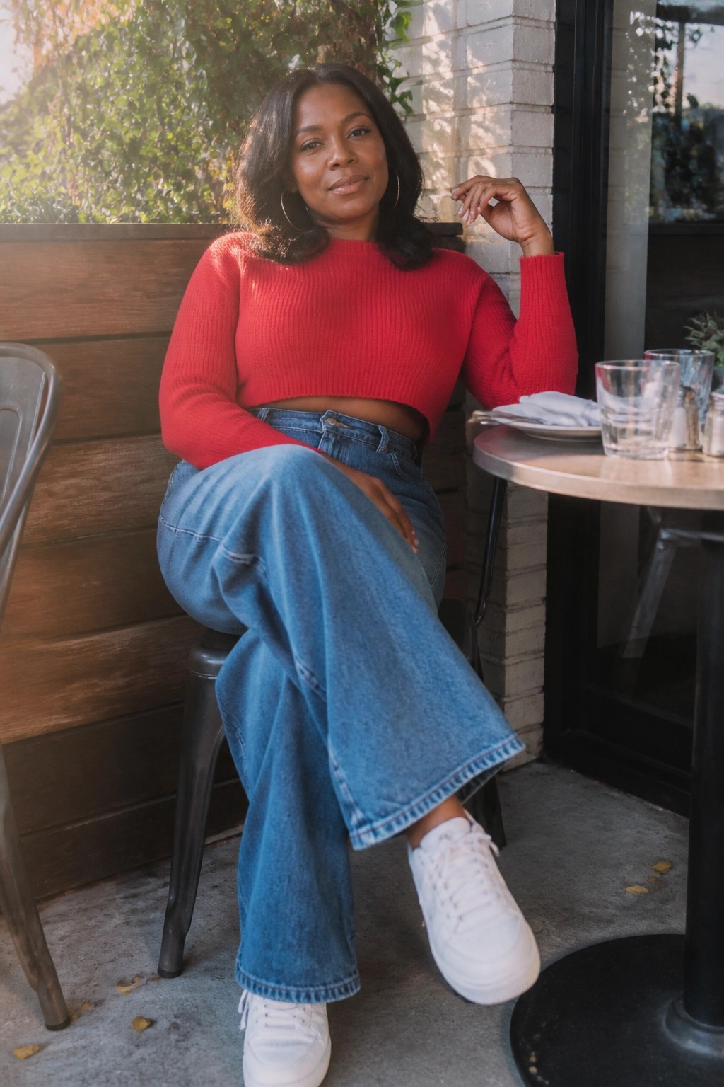 Woman in red cropped sweater and blue jeans seated at sunny outdoor cafe table