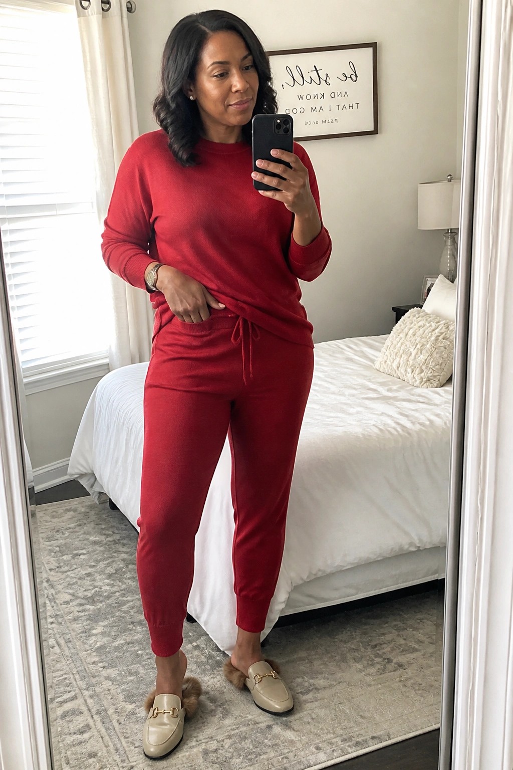 Woman in red knit sweater set holds phone for mirror selfie in bedroom wearing beige loafers