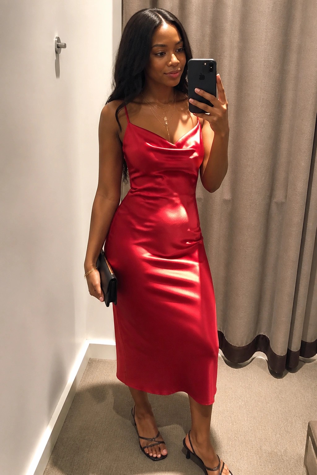 Woman in red satin midi dress taking mirror selfie with phone and clutch.