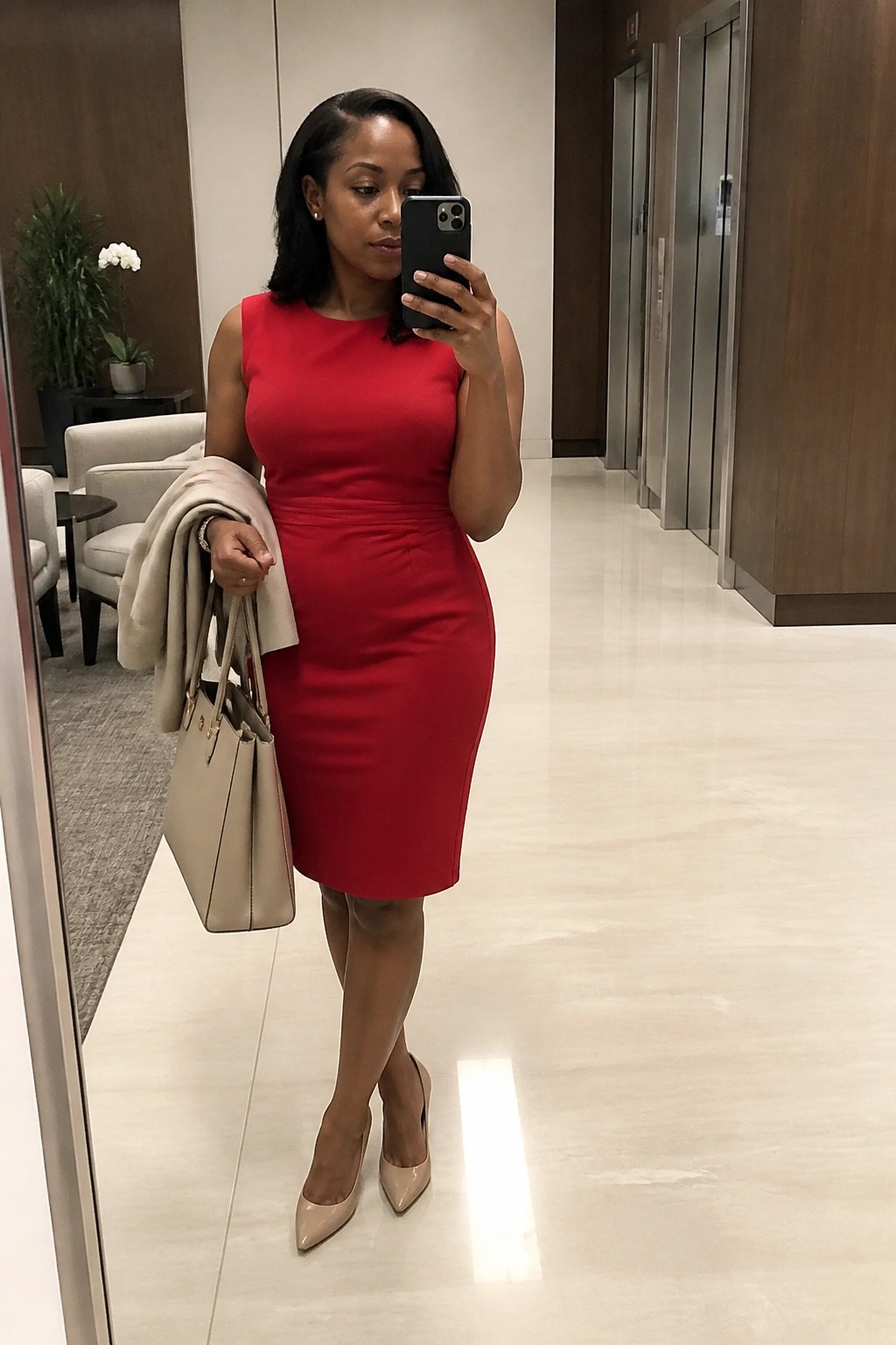 Woman in a fitted sleeveless red sheath dress holding a black phone, with a beige coat draped over one arm, carrying a beige tote bag, and wearing matching nude pointed heels in a hallway setting.