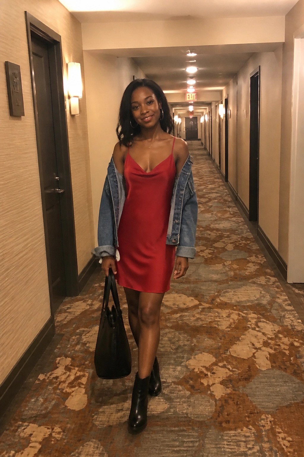 Woman in red slip dress and denim jacket holding black bag in hotel hallway.