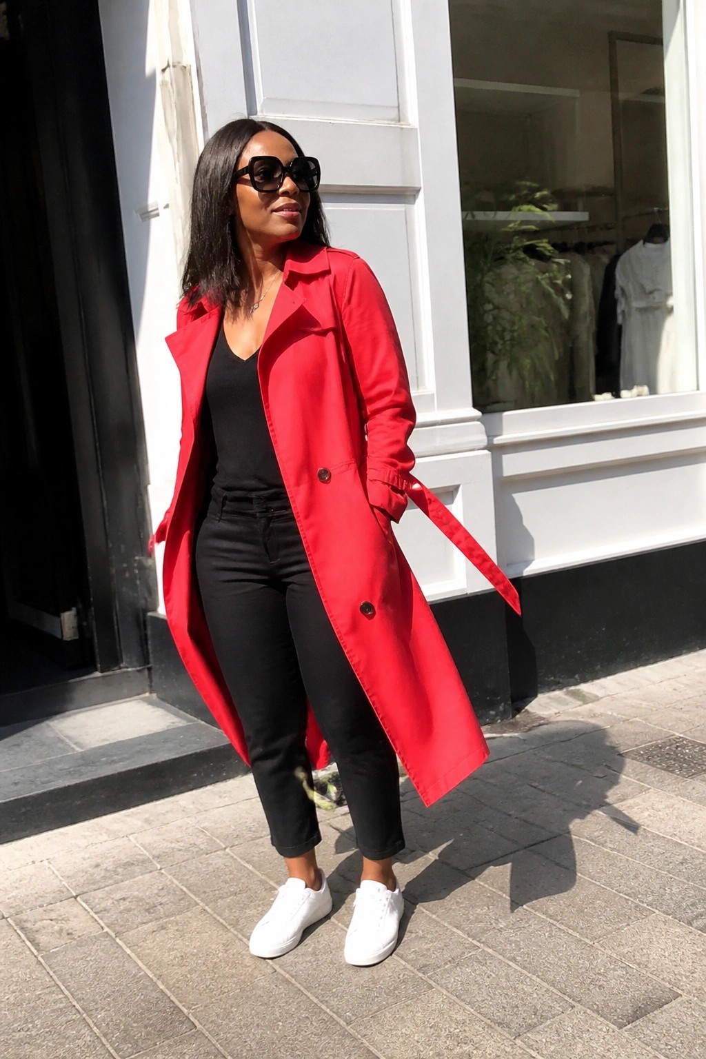 Woman in red trench coat, black top, and white sneakers on sunny sidewalk