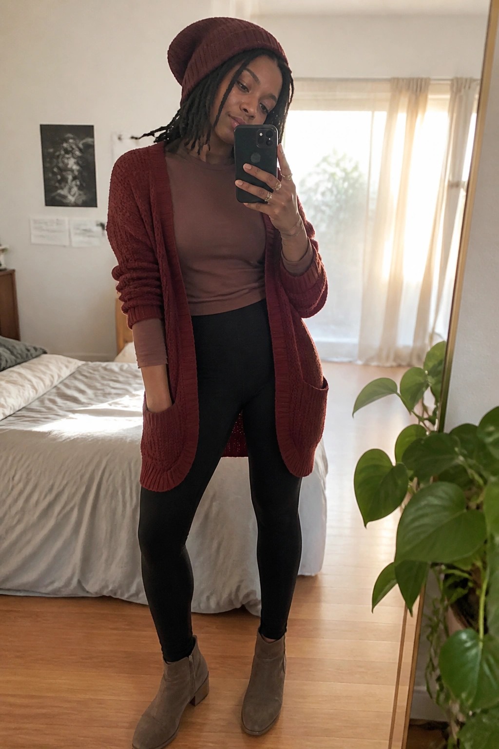 Black woman wearing a long red knit cardigan over a brown top and black leggings with gray suede ankle boots plus a matching red beanie while taking a mirror selfie.
