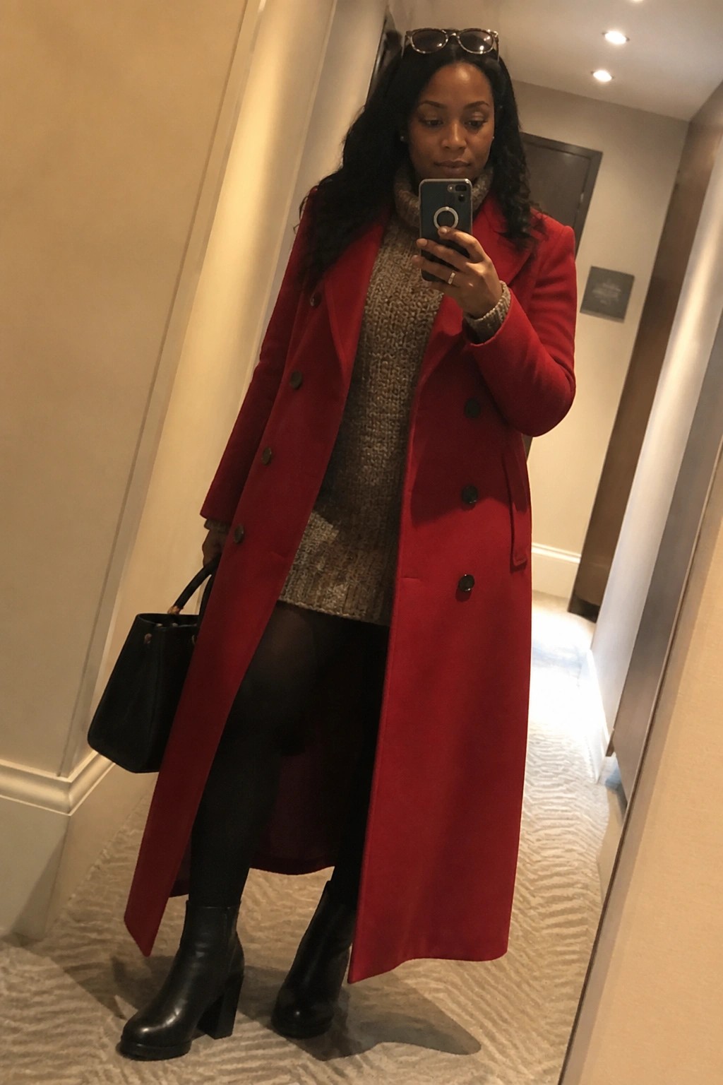 Woman in long red coat and black boots taking mirror selfie in hallway