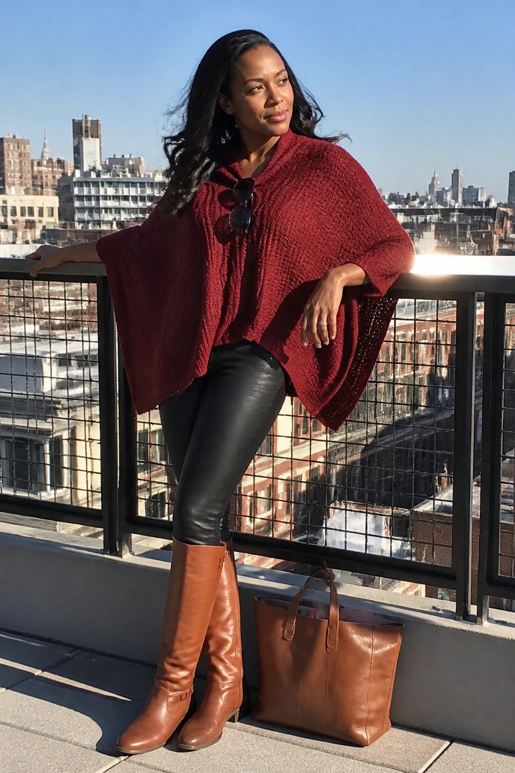 Woman in red poncho and black pants on sunny city balcony with brown boots and bag