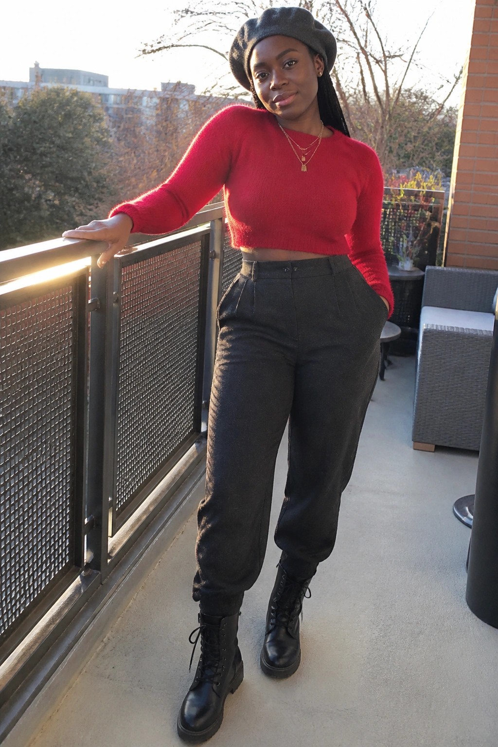 Black woman wearing a cropped red fuzzy sweater, high waisted black textured pants, black combat boots, grey beret, and layered gold necklaces on a balcony.