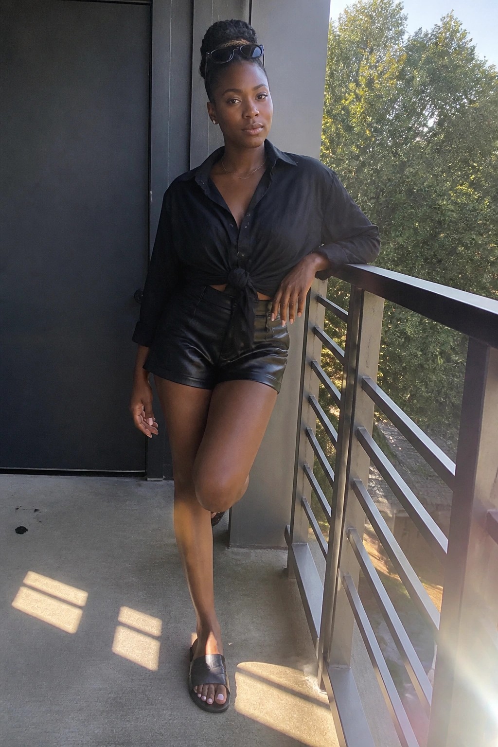 Woman wearing black leather shorts and a black button-up shirt tied at the waist with black slide sandals and sunglasses on her head.