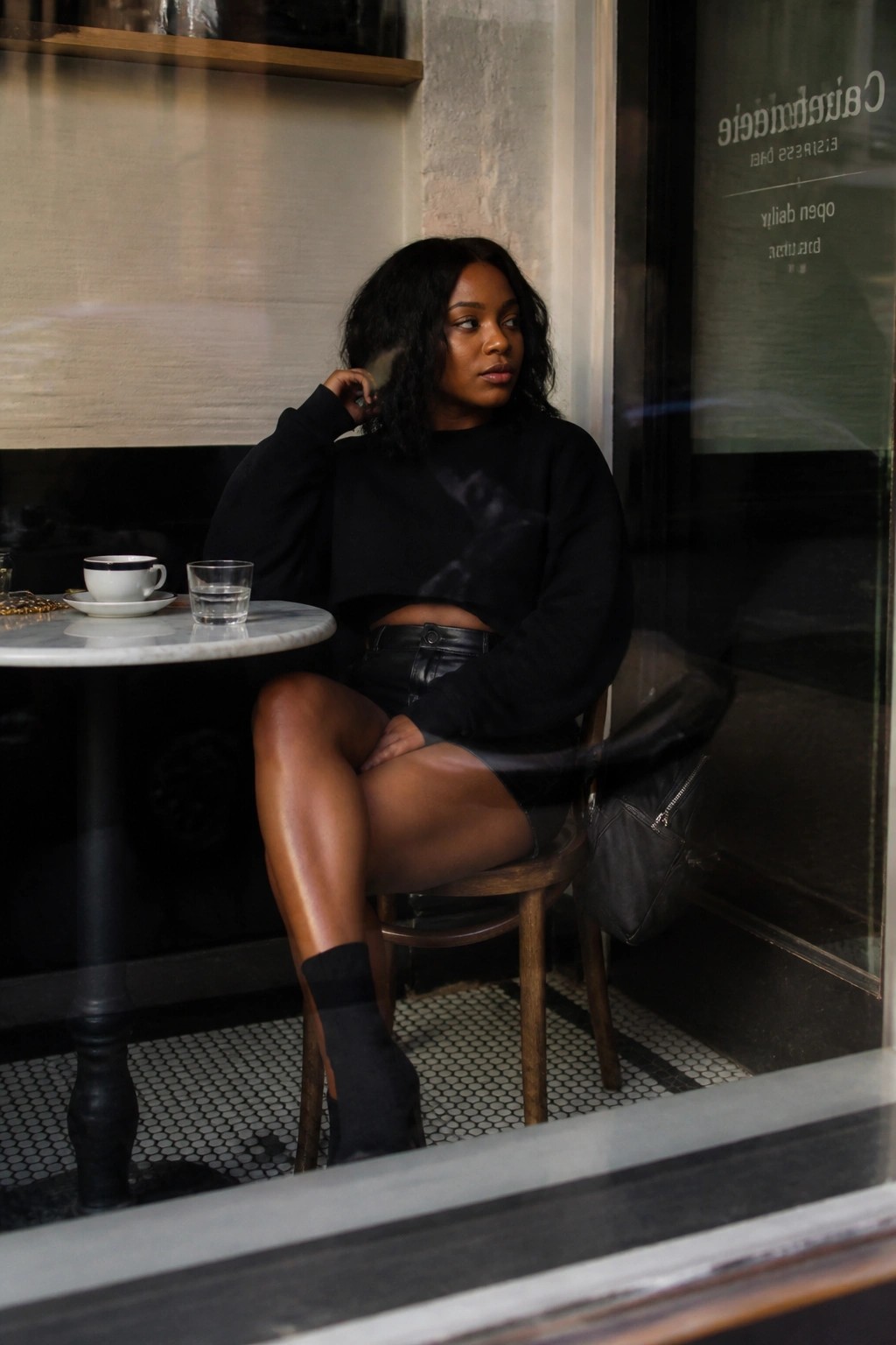 Woman in black cropped sweatshirt and leather skirt seated at marble cafe table
