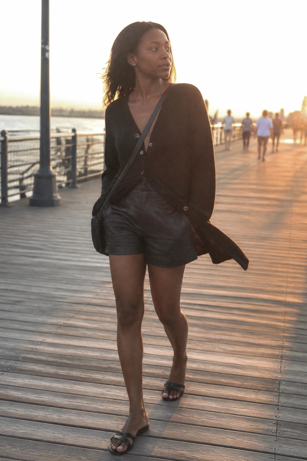 Woman wearing black leather shorts, open black knit cardigan, black crossbody bag, and black slide sandals on a wooden pier.