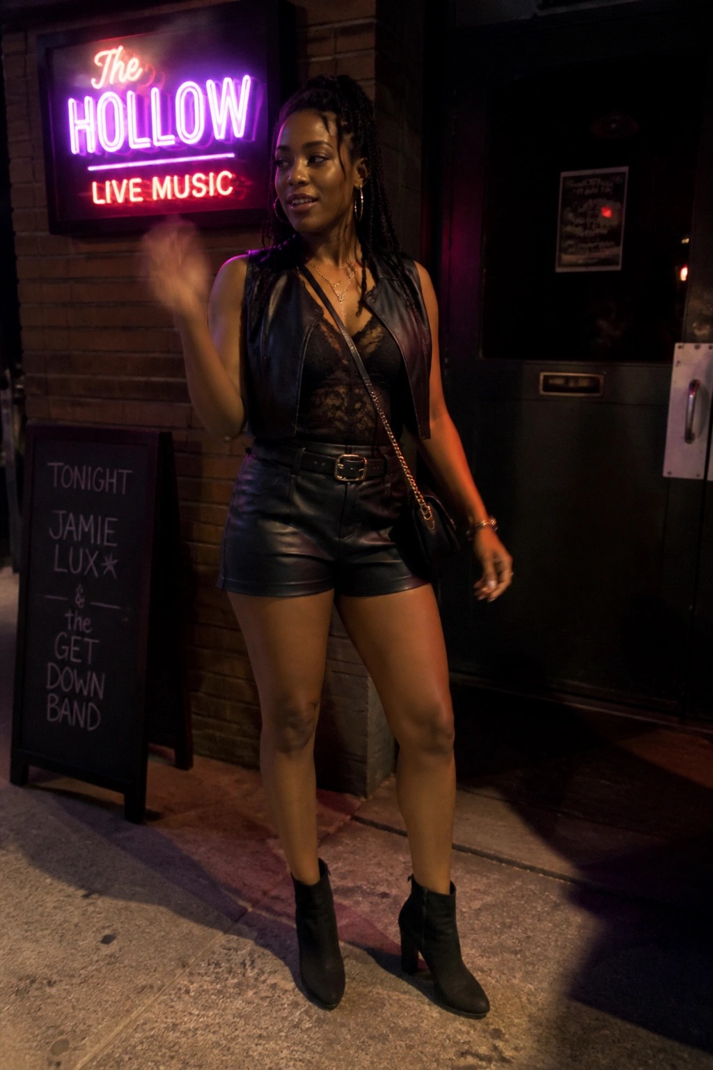 Woman in black leather vest and shorts waves by neon music venue sign at night