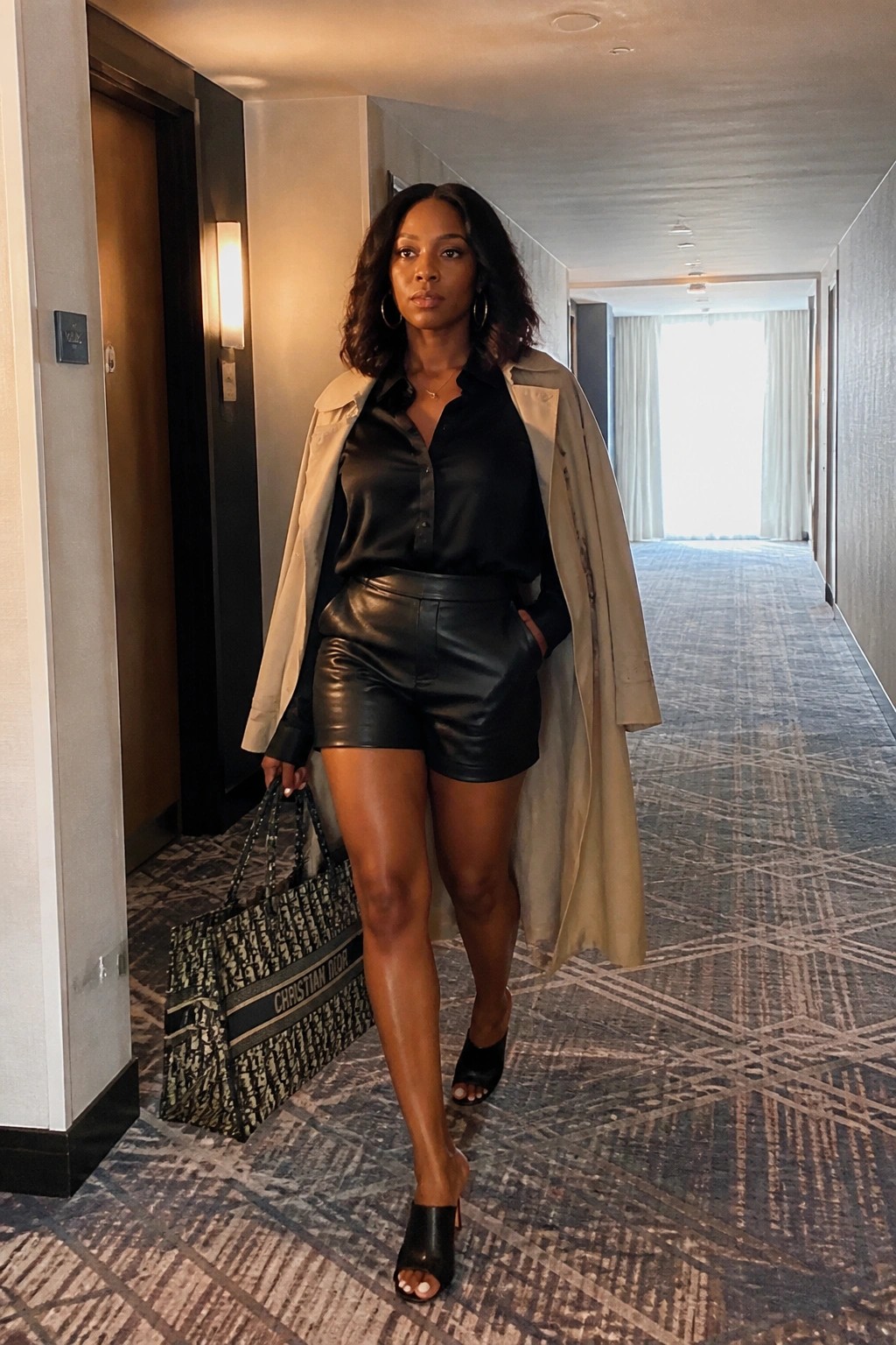Black woman in black leather shorts and matching button up shirt layered with beige trench coat carrying Dior tote bag in hotel hallway.