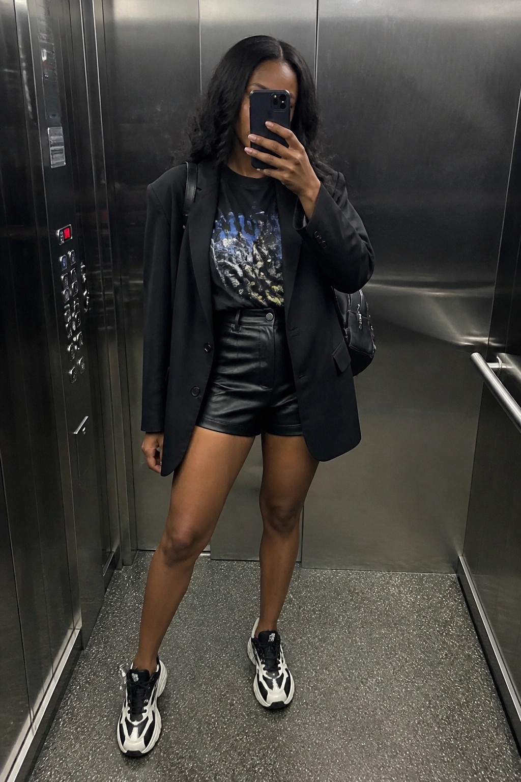 Woman in black blazer and leather shorts taking mirror selfie in elevator