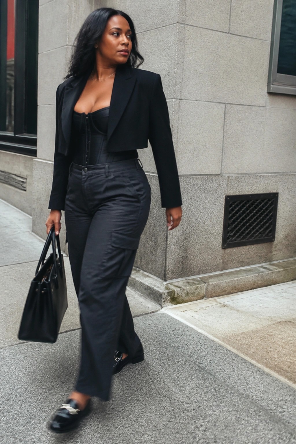 Woman in black cropped blazer, corset top, and cargo pants walks with tote bag.