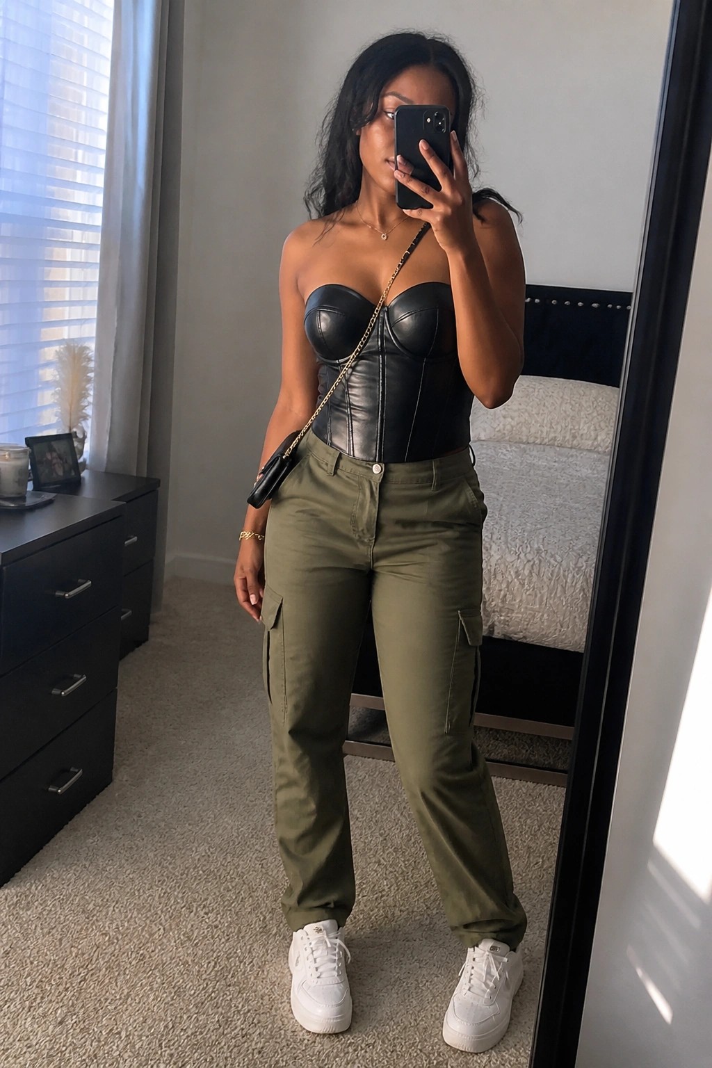 A Black woman in a black leather strapless corset top and olive cargo pants with white sneakers takes a mirror selfie while wearing a gold chain crossbody bag.