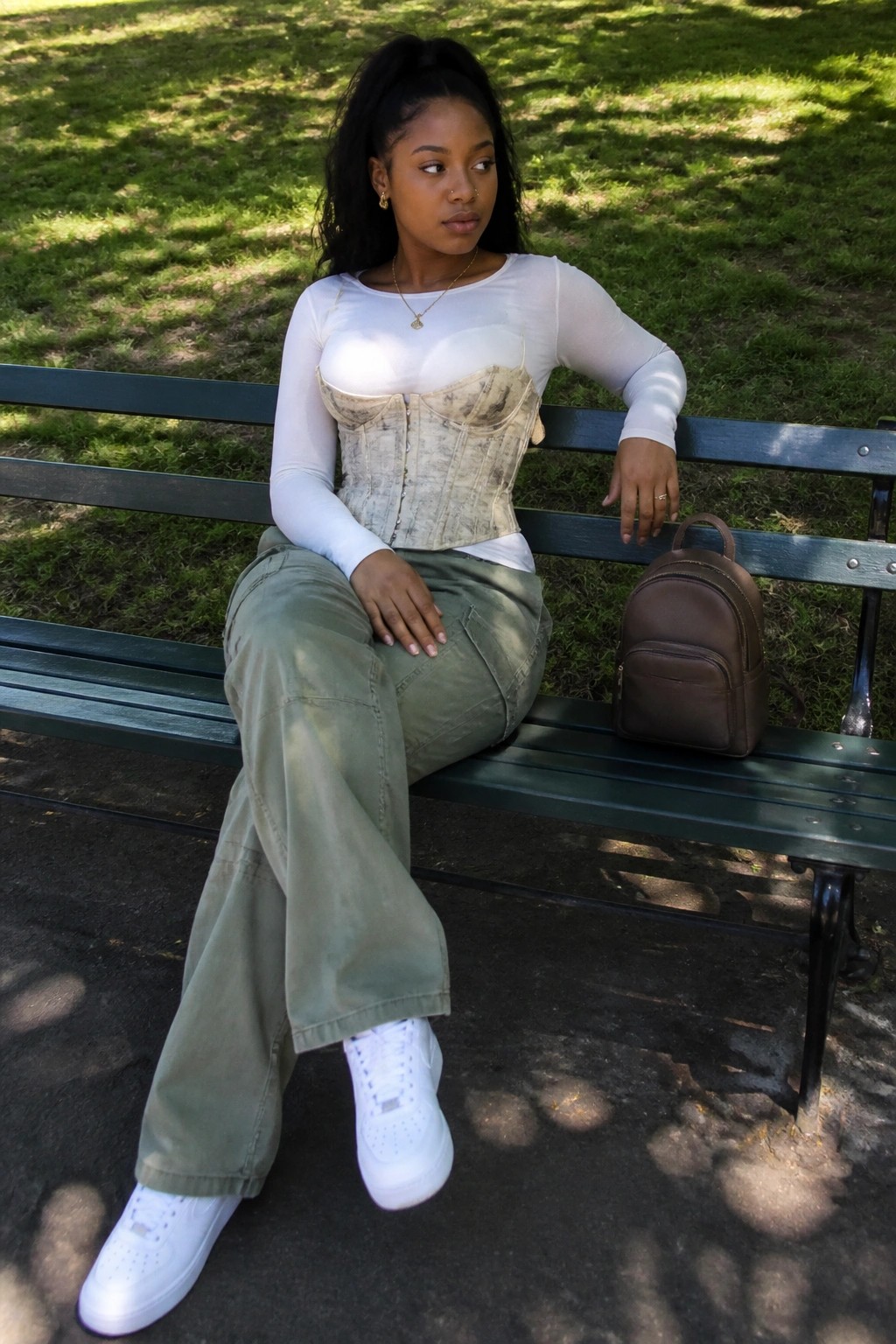 Woman in beige corset and green cargo pants on park bench with backpack.