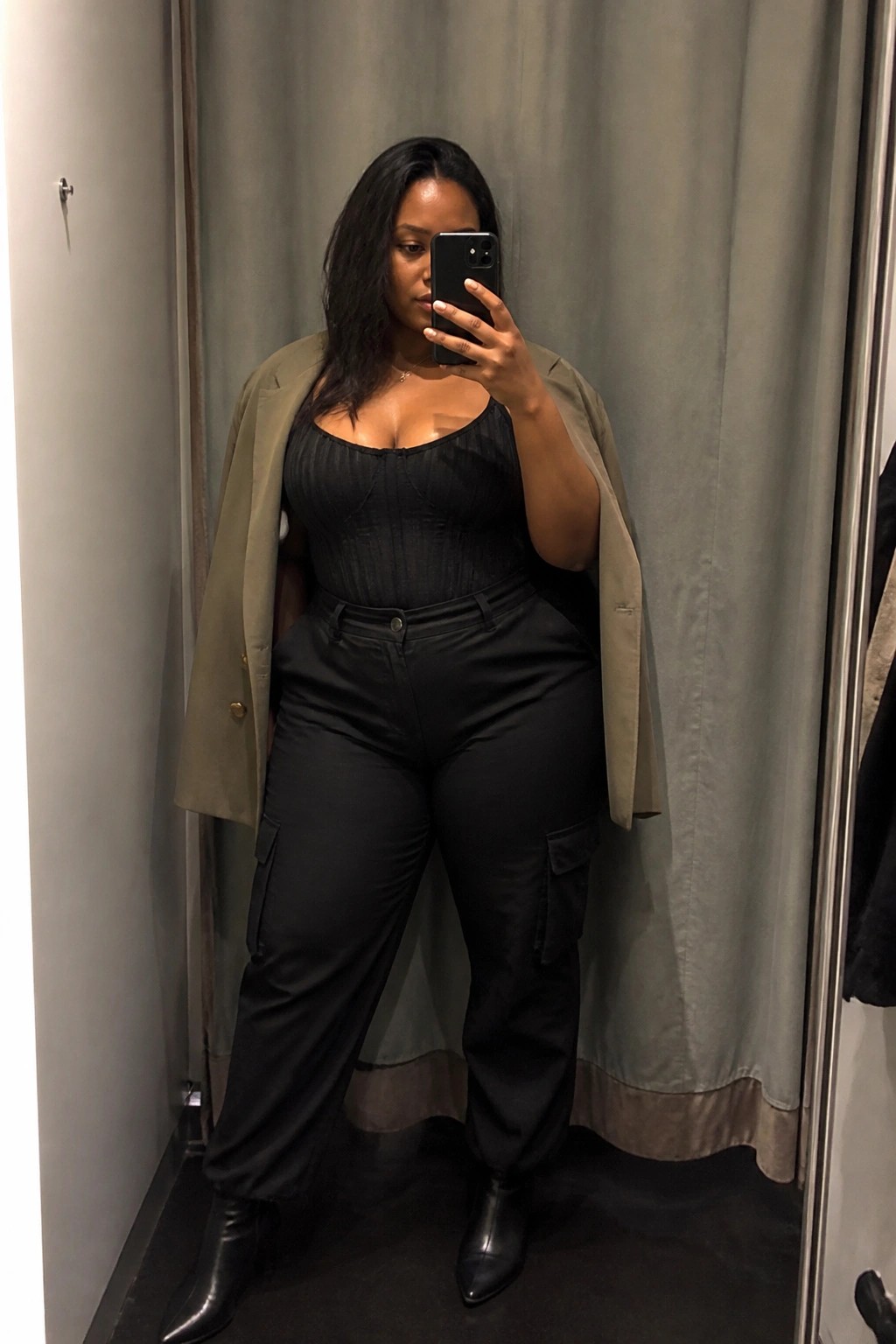 Woman in black corset top, cargo pants, and draped olive blazer taking mirror selfie