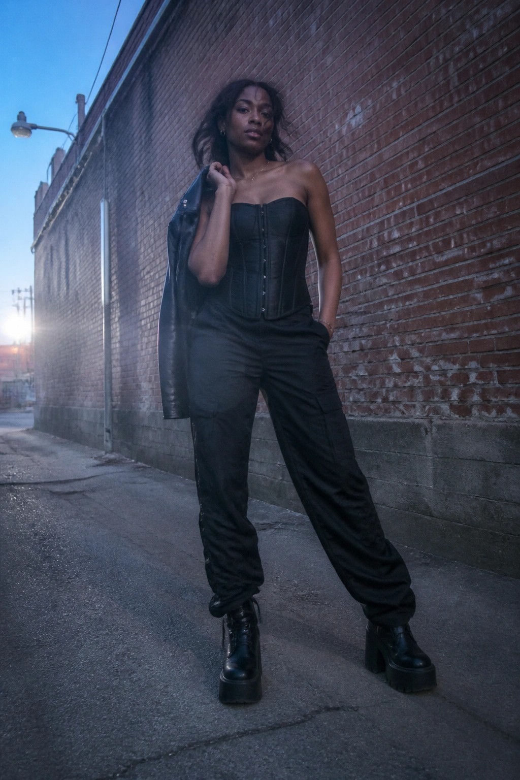 Woman in black corset and cargo pants holds leather jacket in brick alley