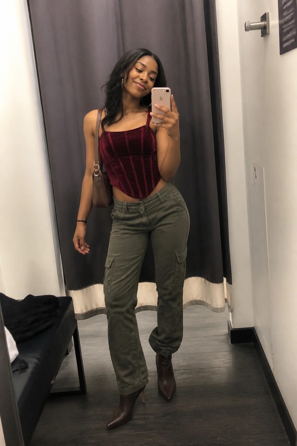 Woman in red velvet corset and green cargo pants taking mirror selfie in fitting room.